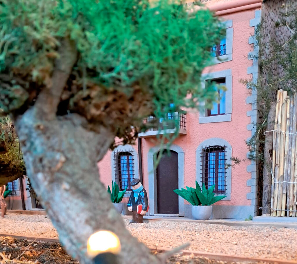 Detall del pessebre de la Fundació Josep Pla, obra de Lluís Brias: una figureta amb un llibre davant la porta d’un edifici, amb un arbre en primer pla.